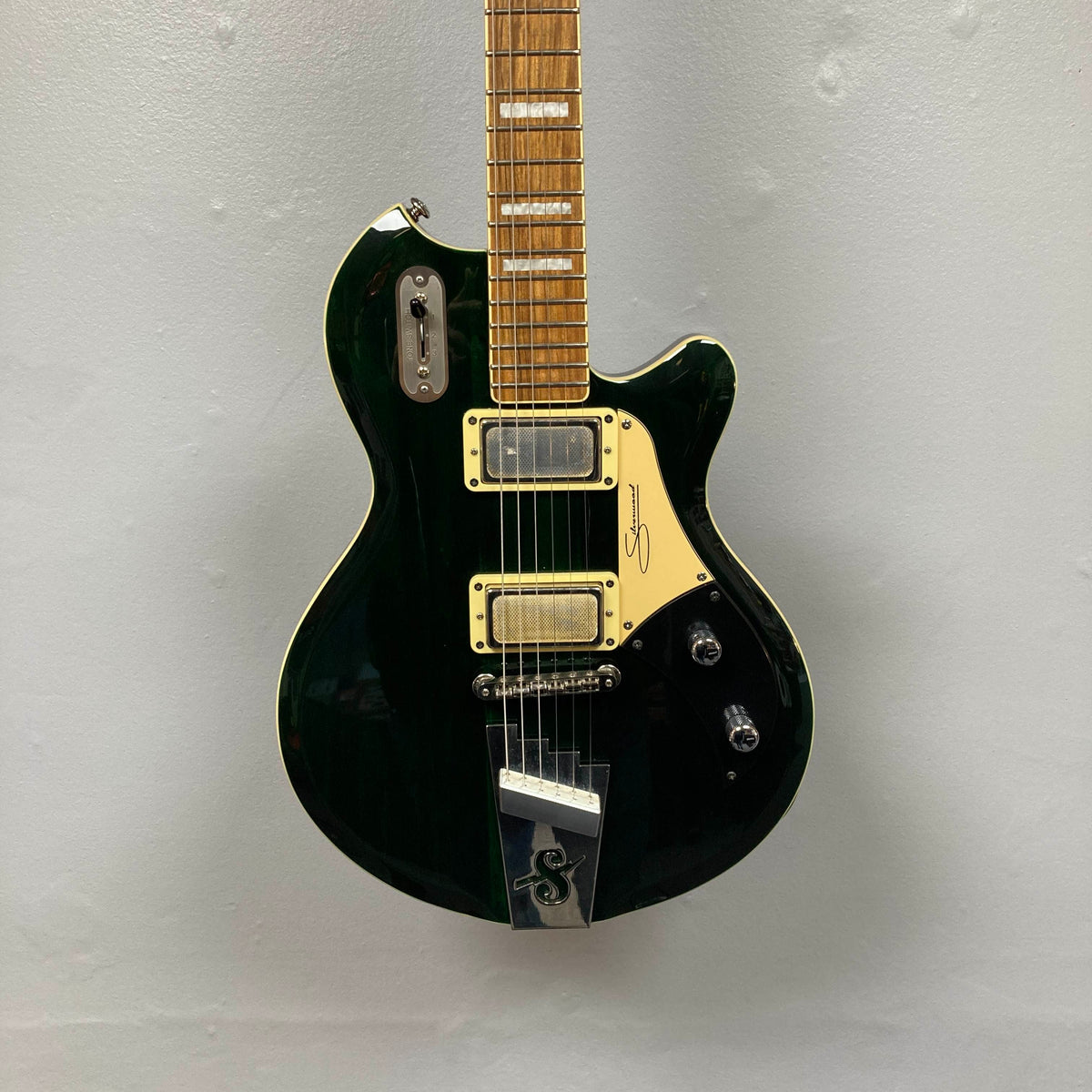 A striking Supro 1296 Silverwood electric guitar in British Racing Green, featuring two Gold Foil pickups, a tremolo bridge, and gold hardware, is mounted against a plain gray wall.