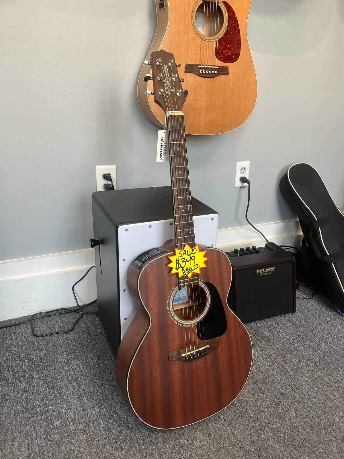 A Takamine GX11ME Taka-Mini NS 3/4 Size Acoustic Guitar w/Case (USED/consign) rests against a white speaker, with another portable guitar, amplifier, and guitar case visible in the background.