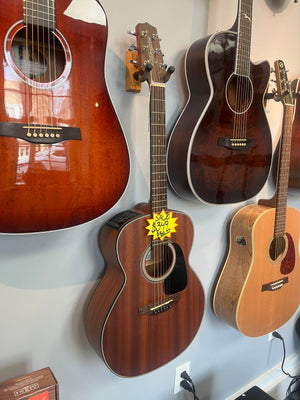 A row of acoustic guitars hangs on a store wall, including a Takamine GX11ME Taka-Mini NS 3/4 Size Acoustic Guitar w/Case USED/consign, marked down from $280 to $229 with a yellow sale sign.