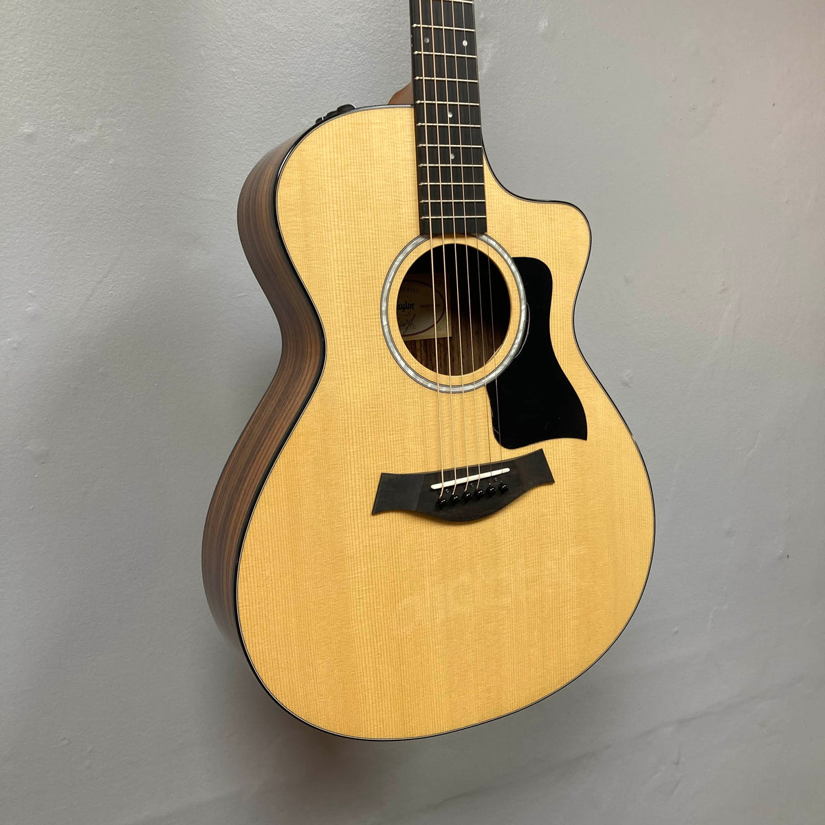 Taylor 212ce Plus w/Gigbag Used Consign by Taylor, featuring a light wood top, cutaway body, and dark wood sides, displayed on a light gray wall.