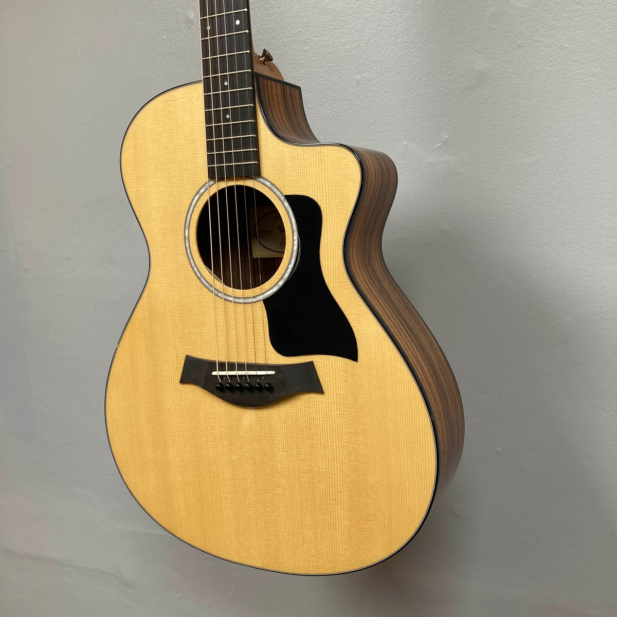 Taylor 212ce Plus w/Gigbag Used Consign by Taylor, featuring a light wood top, dark wood sides, and black pickguard, is displayed hanging on a light gray wall.