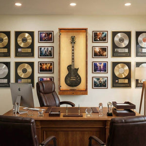A home office with a wooden desk, two leather chairs, and a museum-quality wall display featuring framed records, photos, and The ShowCase™ Deluxe Guitar Display Cabinet by A&S Crafted Products with humidity control.