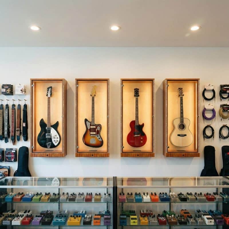 Four guitars are showcased in A&S Crafted Products’ The ShowCase™ Deluxe Lockable Guitar Display Cabinet with LED lighting, mounted above a glass counter stocked with guitar pedals and accessories in the music store.