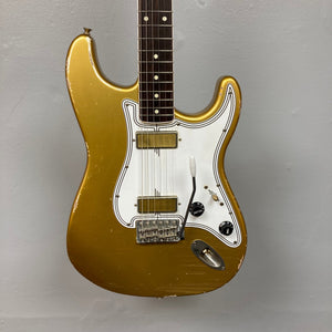 TopHat Stratavarius Aztec Gold Relic electric guitar with lacquered neck, tremolo bridge, and signature detailing, displayed on a wall.