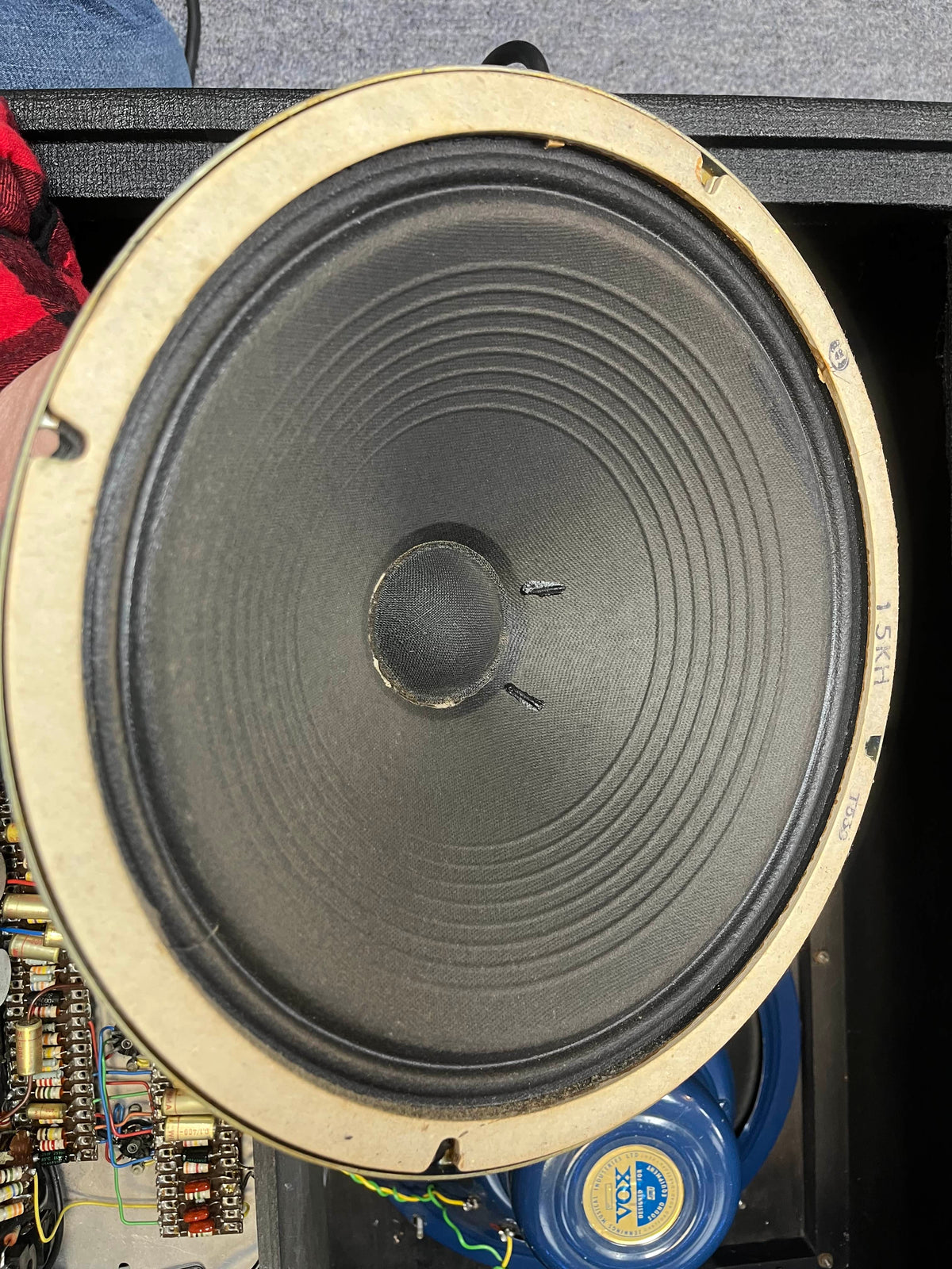 Close-up of a round speaker cone with visible ridges, resembling that of the Vintage 1963 Vox AC30/6 Normal by Vox; mounted in an enclosure with electronic components and wiring partially visible around the edge.