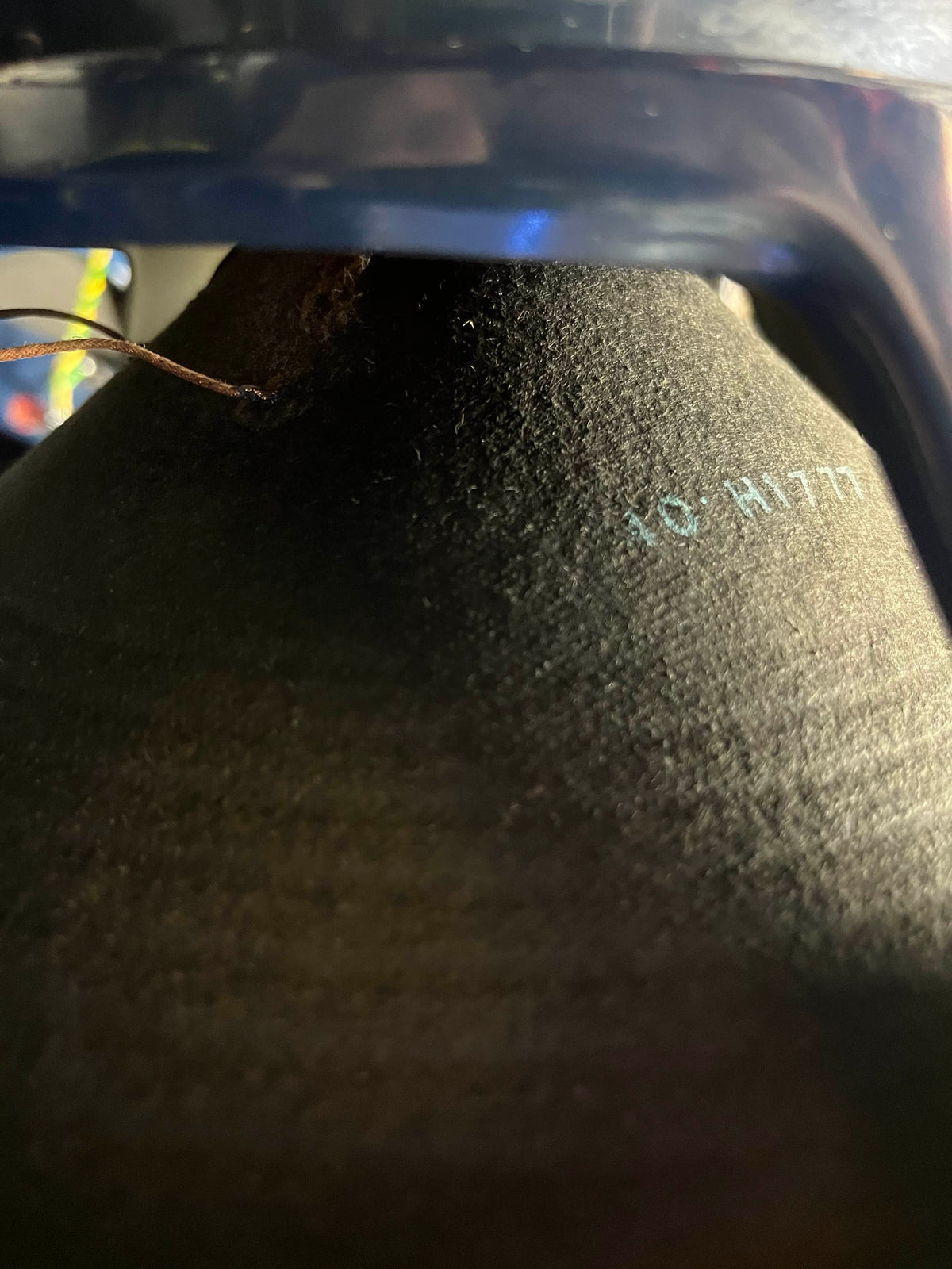 Close-up of a black surface with “16-M1771” in light blue, partly shadowed by a curved dark object—possibly part of a Vintage 1963 Vox AC30/6 Normal by Vox, including the original JMI egg footswitch and flight case.