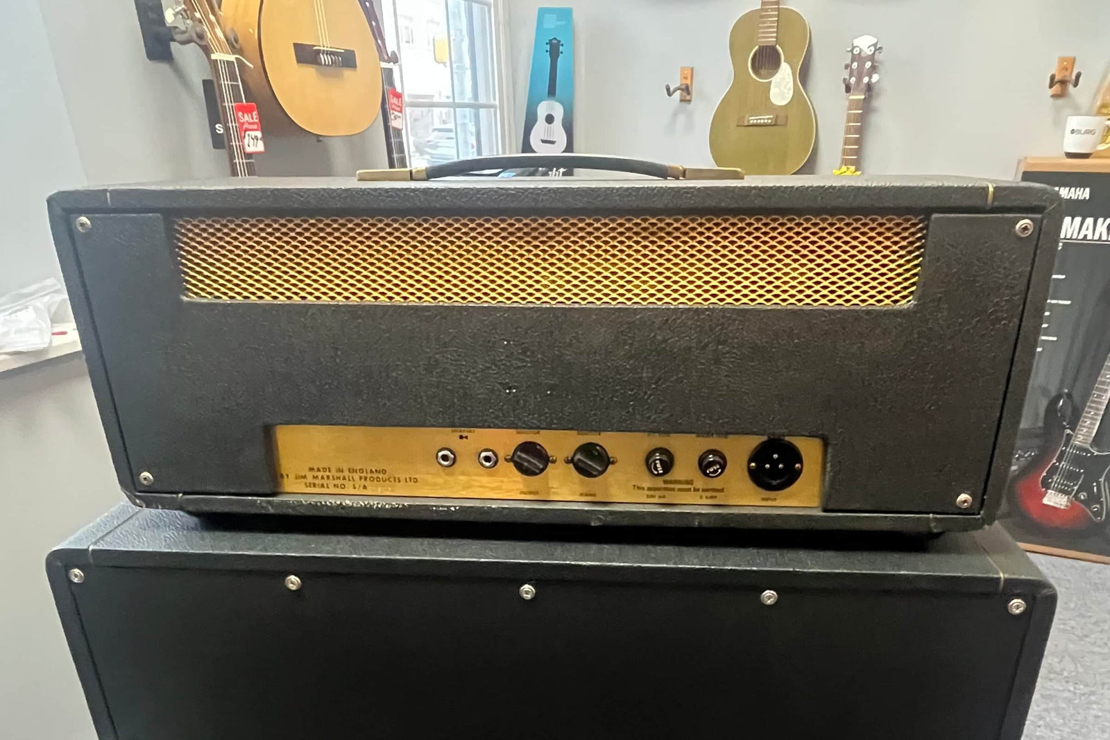 The Marshall 1971 50W Lead Head, JMP Era, with Flight Case rests on a speaker cabinet, showcasing its iconic British sound through its controls. Marshall guitars are displayed in the background.