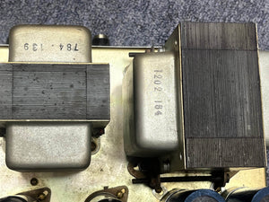 Close-up of two metal-cased transformers with serial numbers, mounted on a chassis—part of a 1971 Marshall 50W Lead Head (JMP Era) from Marshall—shown against a textured gray background.