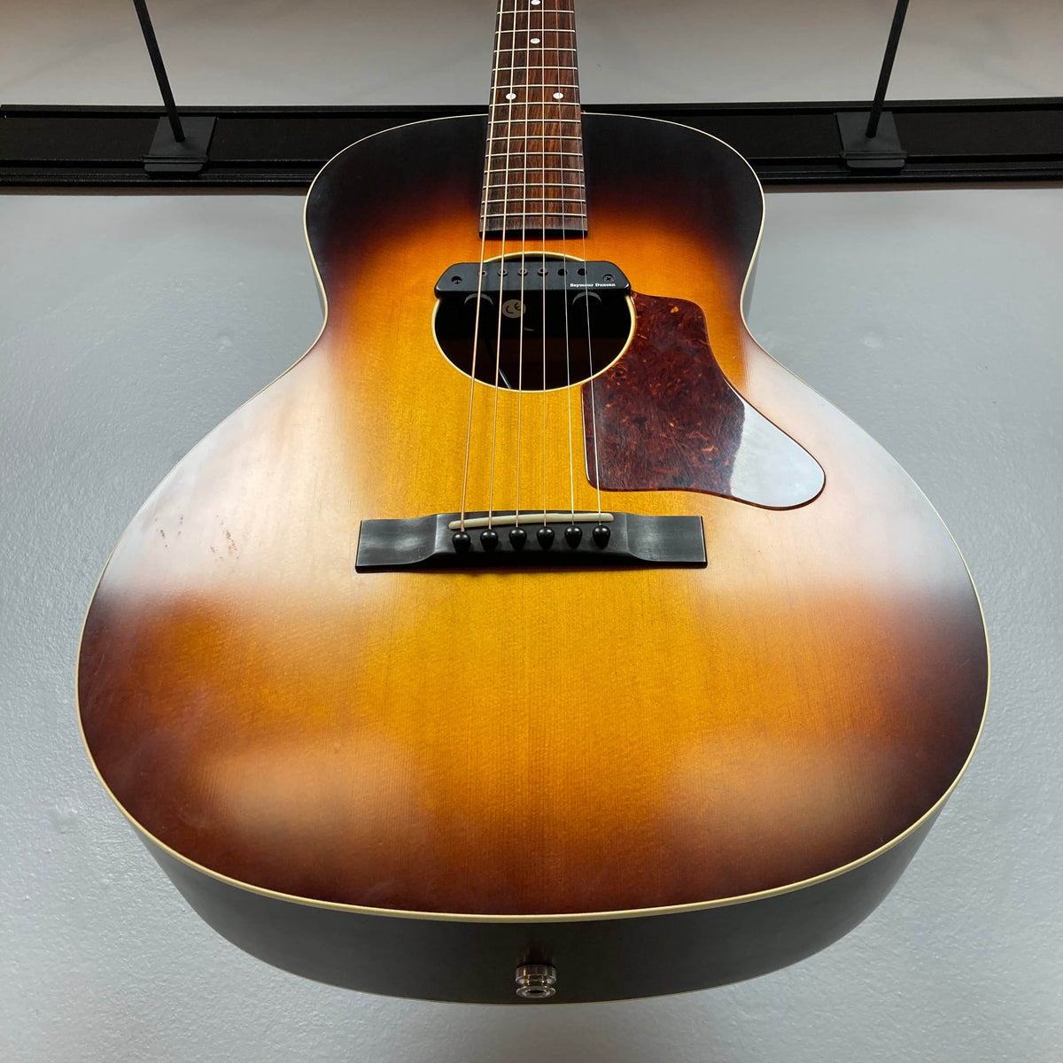A close-up of a Waterloo WL-12 Parlor Acoustic w/Case Used Consign guitar by Waterloo, featuring a sunburst finish, brown pickguard, and six strings, hanging against a plain light-colored wall.