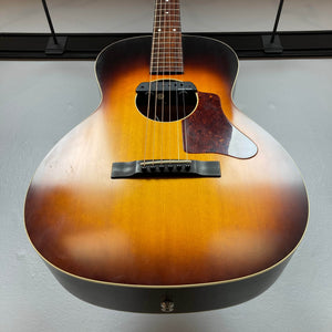 A close-up of a Waterloo WL-12 Parlor Acoustic w/Case Used Consign guitar by Waterloo, featuring a sunburst finish, brown pickguard, and six strings, hanging against a plain light-colored wall.