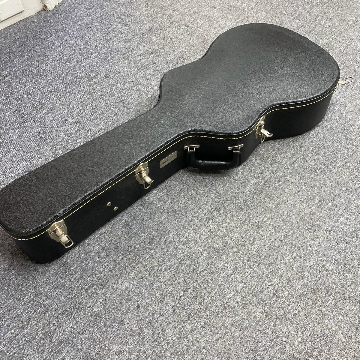 A Waterloo WL-12 Parlor Acoustic used consign guitar case with metal latches and a handle is lying closed on a gray carpeted floor.