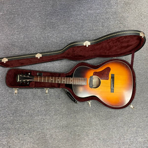A used Waterloo WL-12 Parlor Acoustic guitar with a sunburst finish rests in an open hard-shell case with dark red velvet lining on a gray carpeted floor. Brand: Waterloo. Model: WL-12. Includes case.