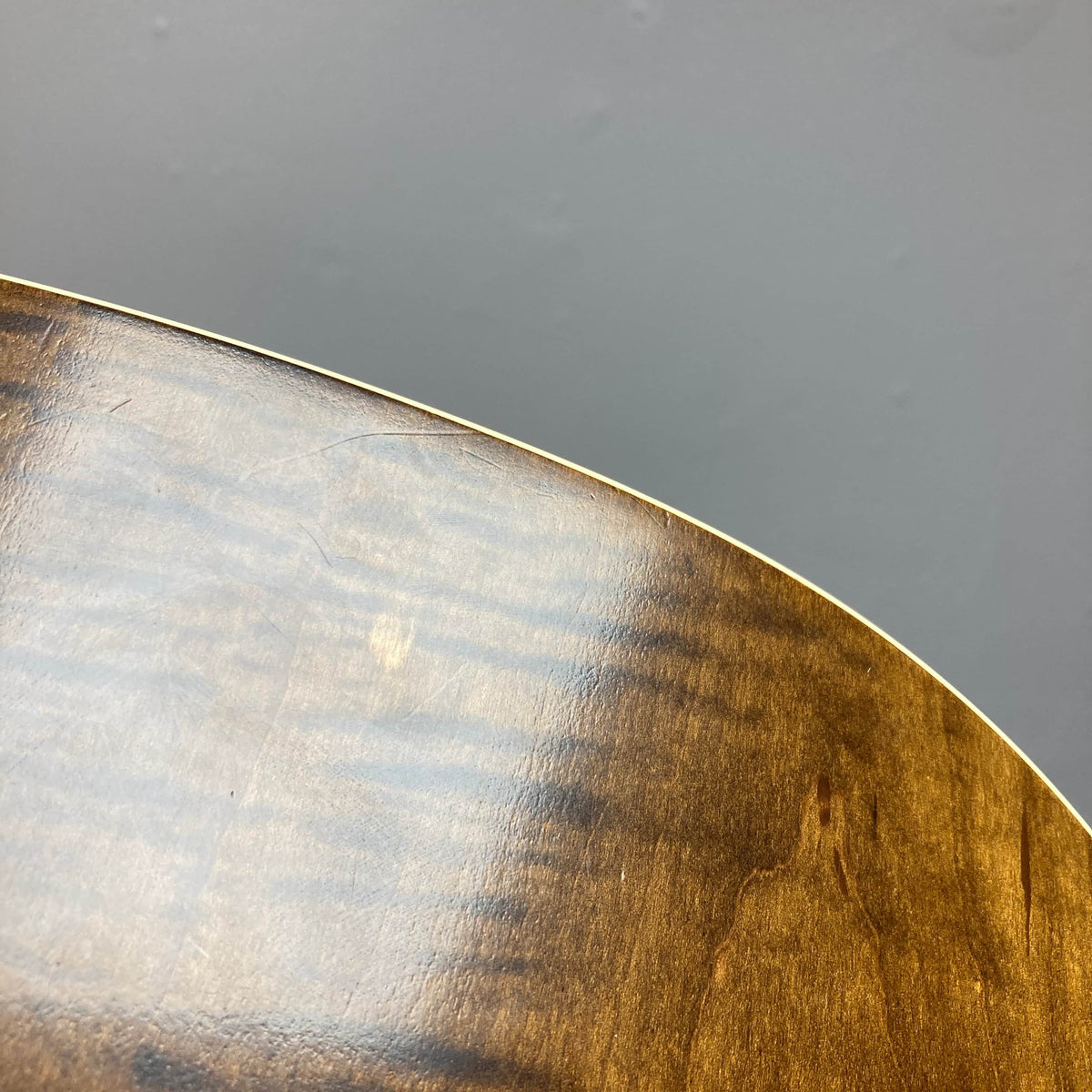 Close-up view of the curved wooden edge and polished surface of a Waterloo WL-12 Parlor Acoustic guitar by Waterloo, with visible wood grain and finish, set against a gray wall.