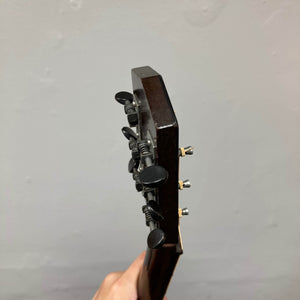 Someone holds the neck of a Waterloo WL-12 Parlor Acoustic guitar, focusing on its headstock and tuning pegs, against a plain light gray background. This is a used consign model from Waterloo, and it comes with a case.