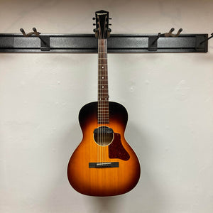 A Waterloo WL-12 Parlor Acoustic w/Case Used Consign hangs on a black wall-mounted rack against a white wall. This vintage-style Waterloo guitar features a sunburst finish, dark headstock, six strings, and brown pickguard.