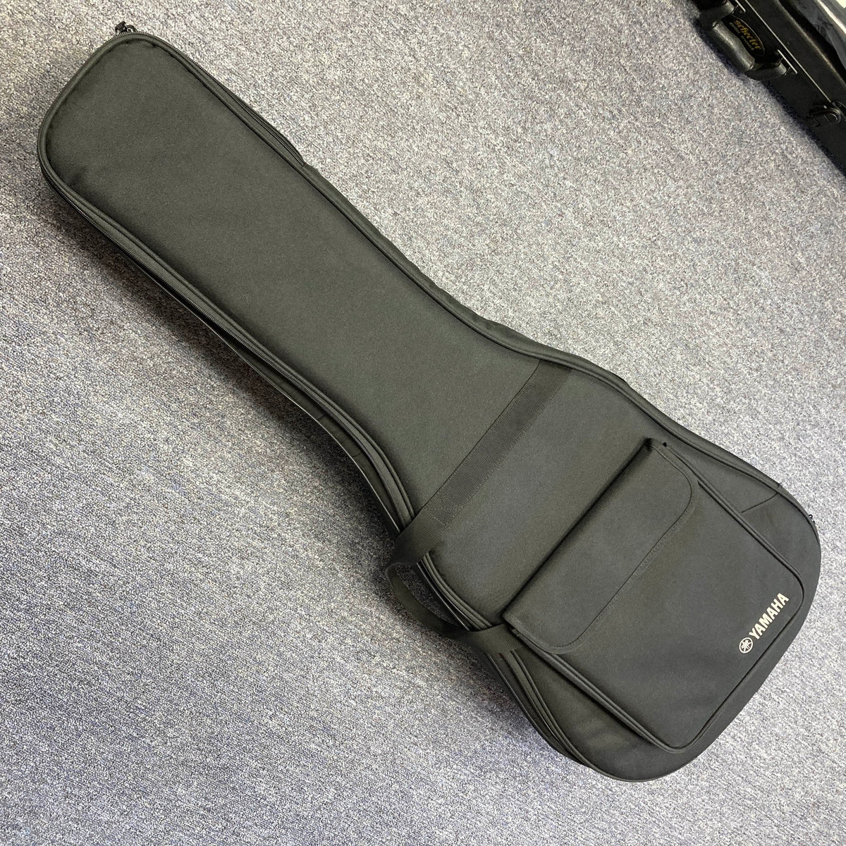 A Yamaha A3M ARE Vintage Natural Acoustic Guitar by Yamaha, featuring a solid Sitka spruce top and SRT2 pickup system, along with its included hard bag, is resting on a gray carpeted floor. The hard bag offers a convenient front pocket for storing accessories.