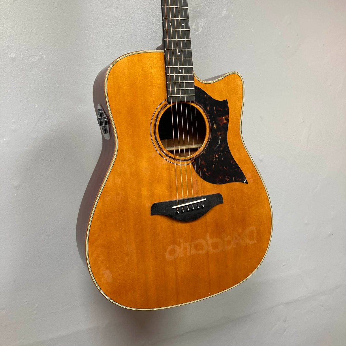 A Yamaha A3M ARE Vintage Natural Acoustic Guitar hangs on the wall, showcasing its cutaway body, tortoiseshell pickguard, and a reflection of D'Addario branding on the finish. This used model features solid mahogany construction and comes equipped with Yamaha’s advanced SRT2 pickup system for exceptional amplified tone; includes a hard bag.