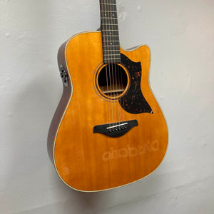A Yamaha A3M ARE Vintage Natural Acoustic Guitar hangs on the wall, showcasing its cutaway body, tortoiseshell pickguard, and a reflection of D'Addario branding on the finish. This used model features solid mahogany construction and comes equipped with Yamaha’s advanced SRT2 pickup system for exceptional amplified tone; includes a hard bag.