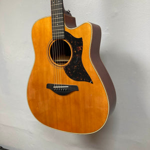 Yamaha A3M ARE Vintage Natural Acoustic Guitar by Yamaha, featuring a cutaway body, solid mahogany construction, advanced SRT2 pickup system, and black pickguard, is hanging on a light gray wall. This used guitar comes with a hard bag and showcases a beautiful vintage natural wood finish.