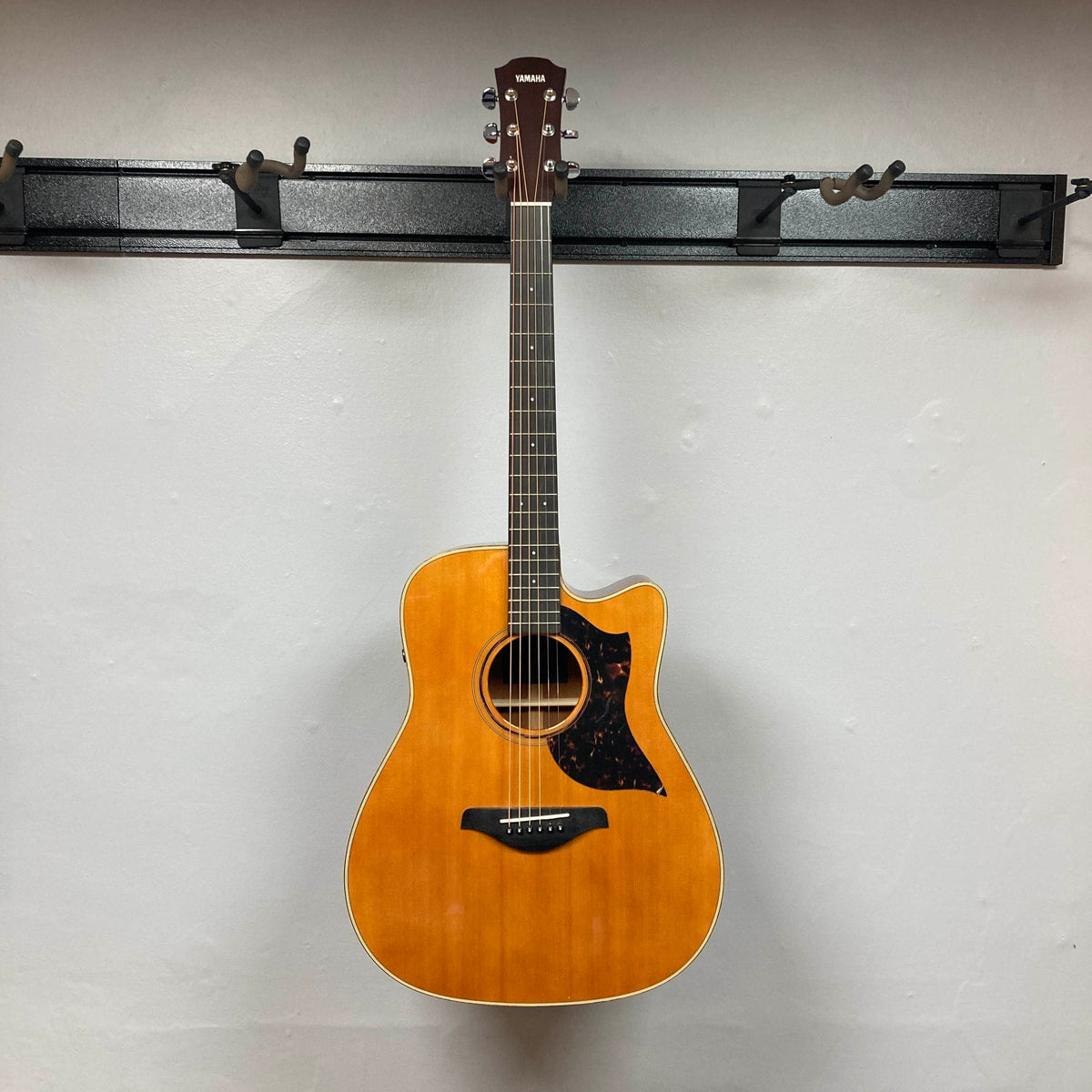 A Yamaha A3M ARE Vintage Natural Acoustic Guitar by Yamaha, featuring a cutaway body and six strings, hangs on a black wall-mounted rack against a plain white wall, highlighting its elegant design and advanced SRT2 pickup system.