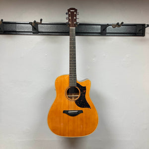 A Yamaha A3M ARE Vintage Natural Acoustic Guitar by Yamaha, featuring a cutaway body and six strings, hangs on a black wall-mounted rack against a plain white wall, highlighting its elegant design and advanced SRT2 pickup system.
