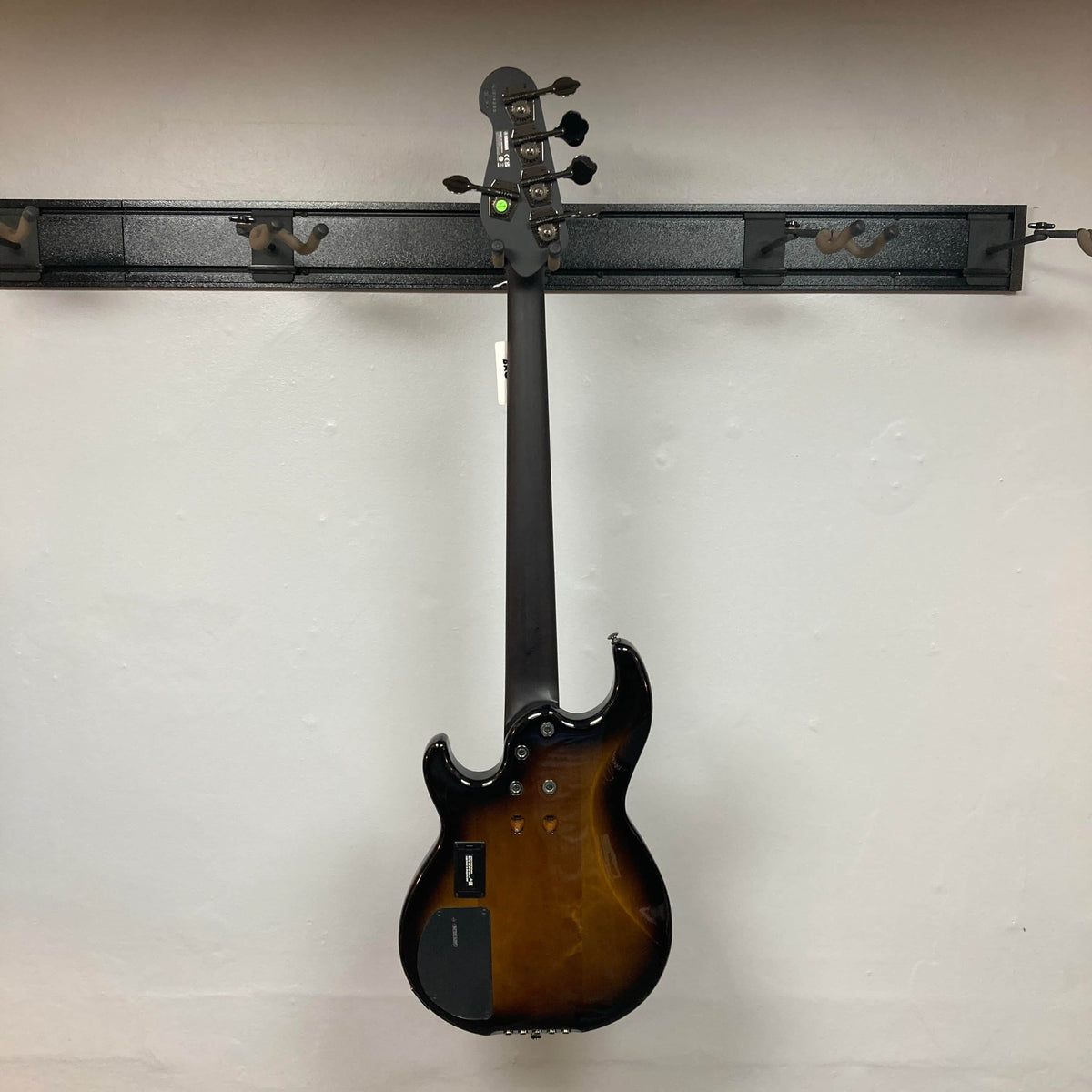 A Yamaha BB735A Electric 5-String Bass Coffee Sunburst (Blem Discount) by Yamaha hangs on a black wall-mounted rack, shown from the back with a dark brown neck, tuning pegs, control plates, and a smooth white wall background.