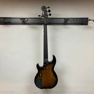 A Yamaha BB735A Electric 5-String Bass Coffee Sunburst (Blem Discount) by Yamaha hangs on a black wall-mounted rack, shown from the back with a dark brown neck, tuning pegs, control plates, and a smooth white wall background.