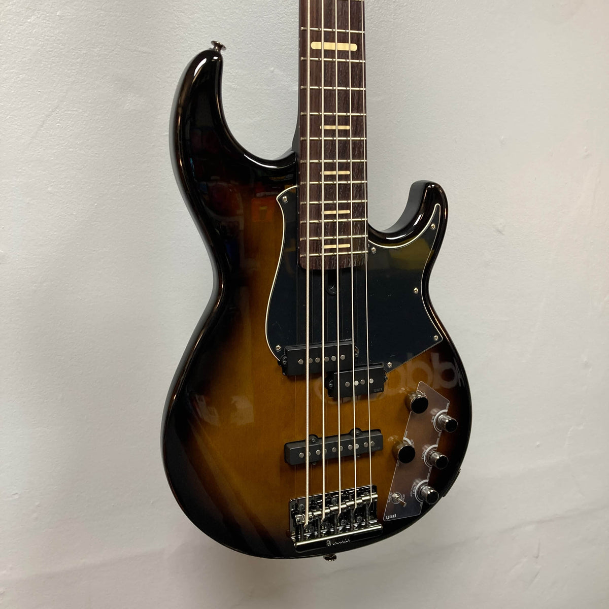 A Yamaha BB735A Electric 5-String Bass in Coffee Sunburst (Blem Discount) with two pickups, multiple control knobs, and a black pickguard hangs against a plain white wall.