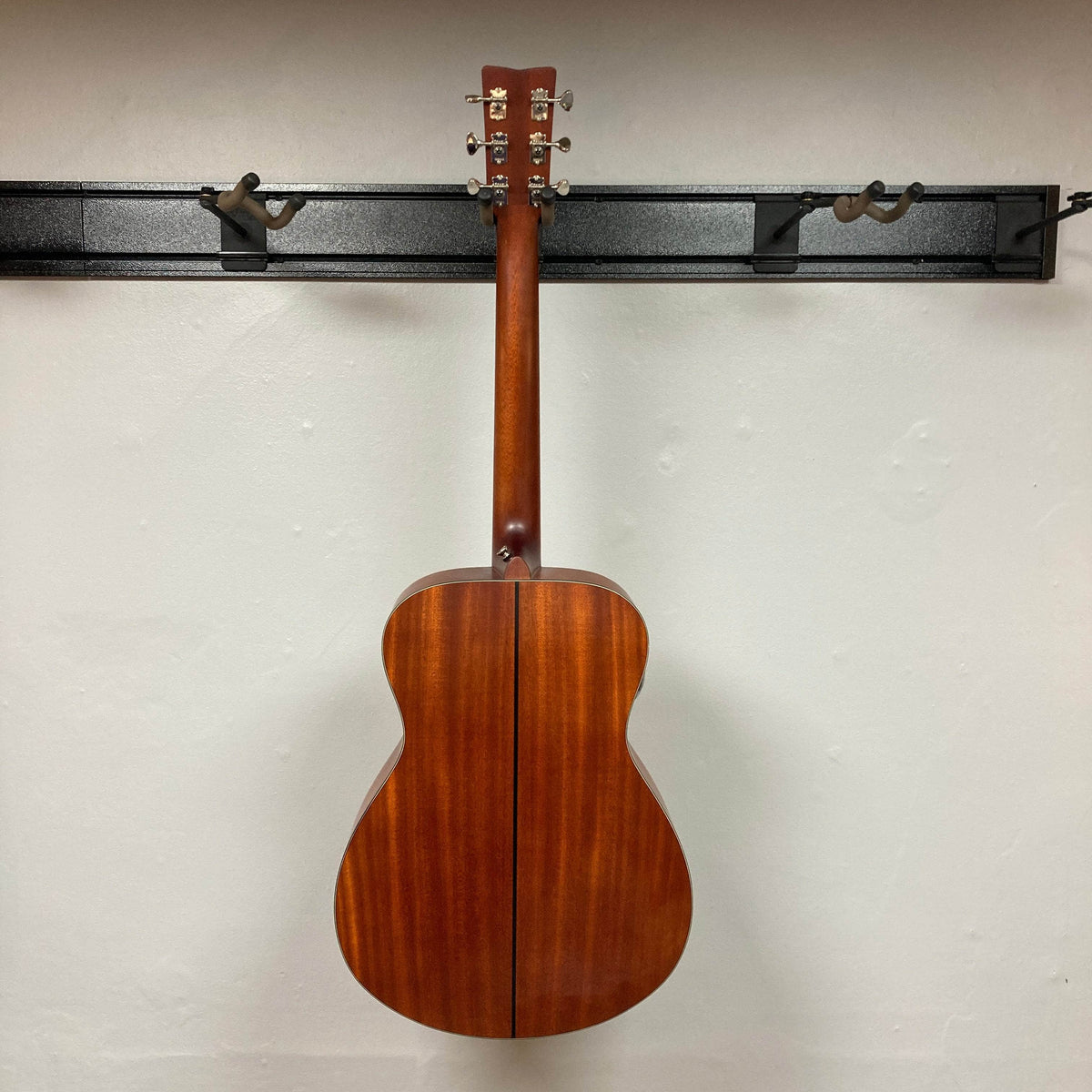 A Yamaha FSX5 Satin Red Label Natural acoustic electric guitar, made in Japan and sold with a case, hangs on a black wall rack, showing its back and neck against a plain white wall.
