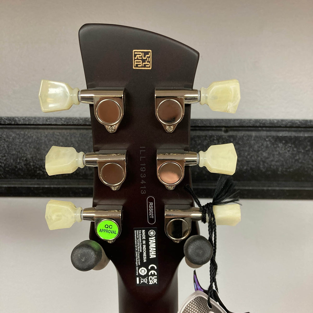 Close-up of the Yamaha Revstar Standard RSS02T Vintage White headstock, showing six tuning pegs, manufacturer labels, serial number, and a green “QC Approved” sticker. A tag hangs from one peg. Includes gigbag. Brand: Yamaha.