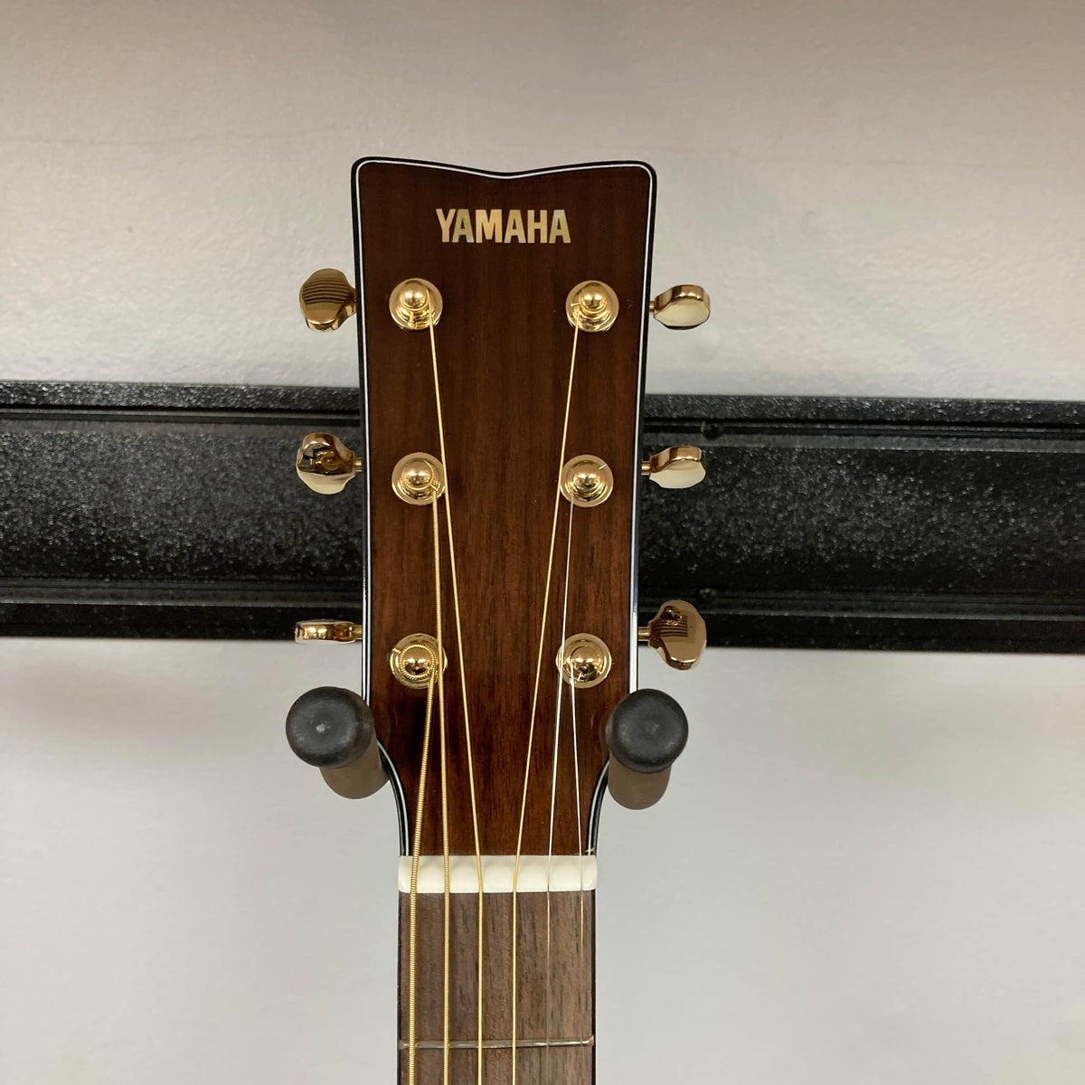 The Yamaha Storia III Solid Mahogany Top Concert guitar's headstock with gold tuning pegs is displayed on a wall-mounted holder.