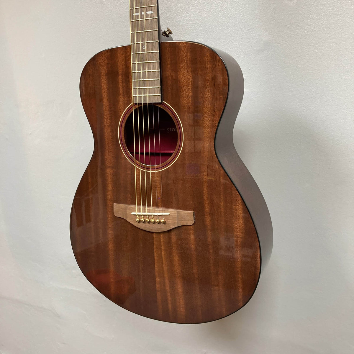 The Yamaha Storia III Solid Mahogany Top Concert guitar by Yamaha, featuring a brown glossy finish, hangs on a white wall.