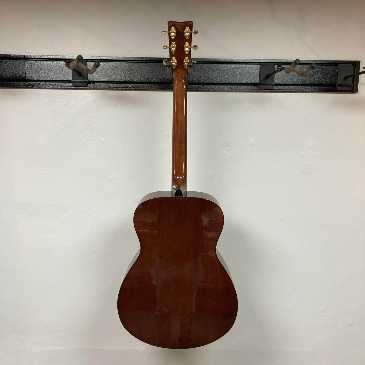 A Yamaha Storia III Solid Mahogany Top Concert guitar by Yamaha hangs back-facing on a black wall-mounted rack against a white wall.