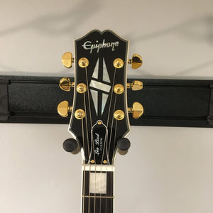 Headstock of 2022 Epiphone Les Paul Custom Ebony with pearl inlay, gold tuners, featuring Gibson pickups, on wall-mount guitar hanger.