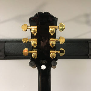 Back of 2022 Epiphone Les Paul Custom Ebony showing gold tuning machines, inspection sticker, and Hand-crafted in China label, mounted on a wall hanger.