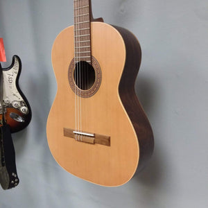 Ortega Nylon String Guitar R189SN-25TH, displayed on a wall, featuring a solid American cedar top and walnut fretboard. Comes with a 25th Anniversary gig bag.